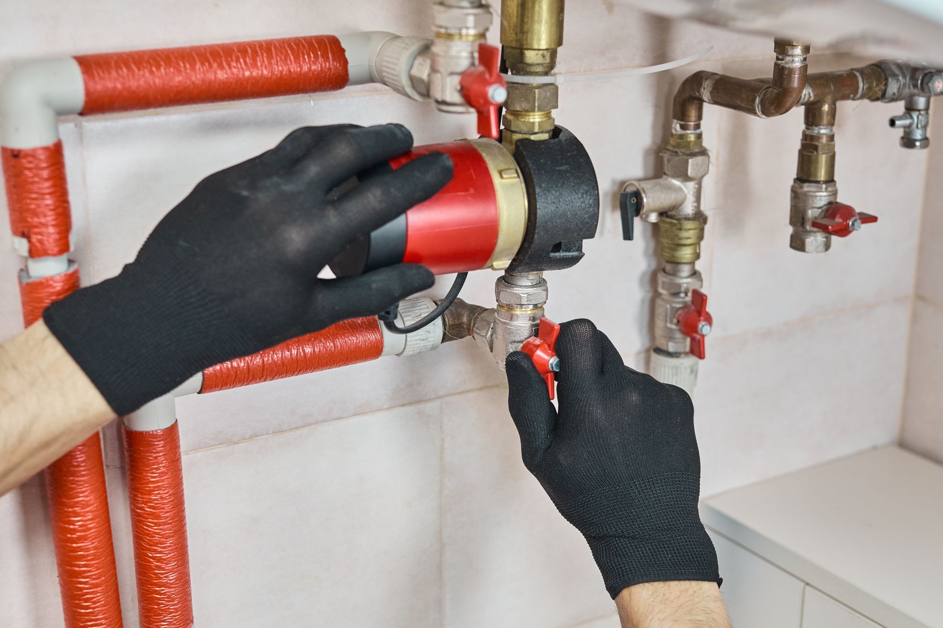 Gloved hands adjusting a red valve on plumbing pipes under a sink