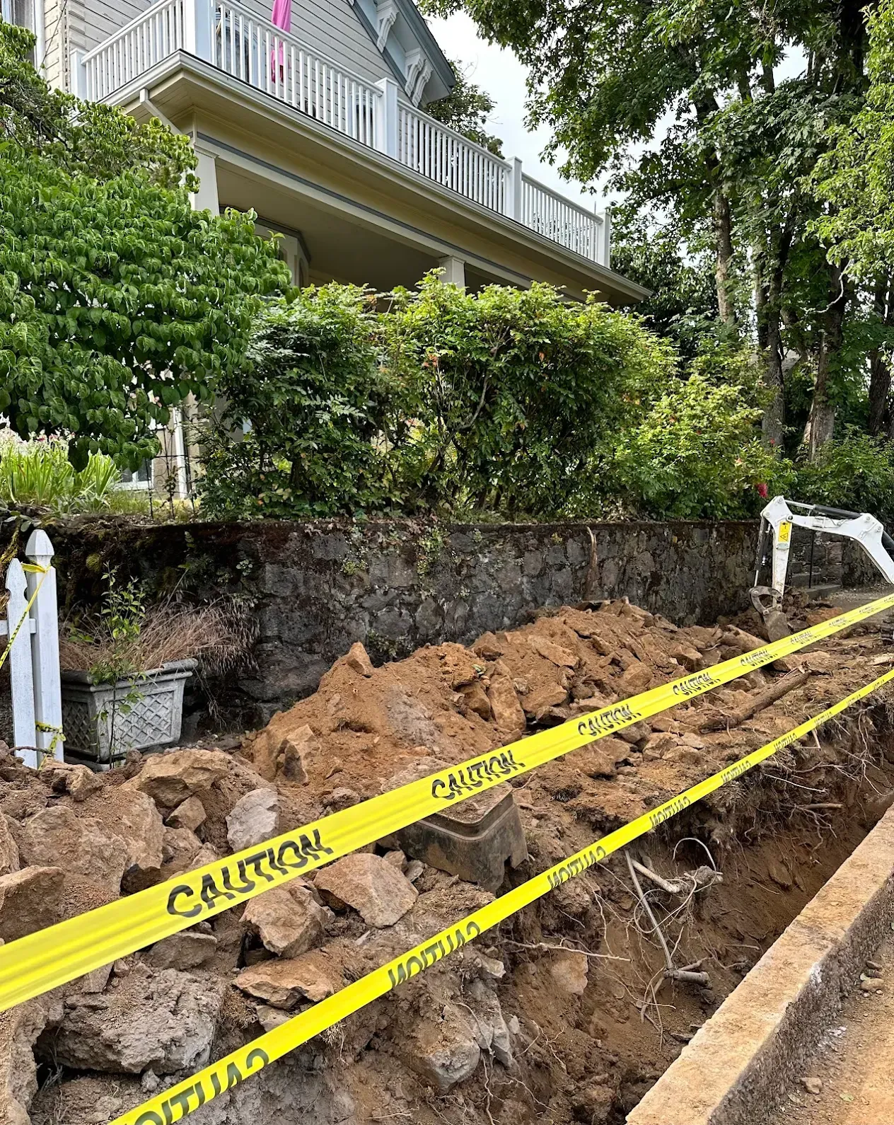 Caution tape across a dirt excavation beside a house and hedge, with a balcony overhead