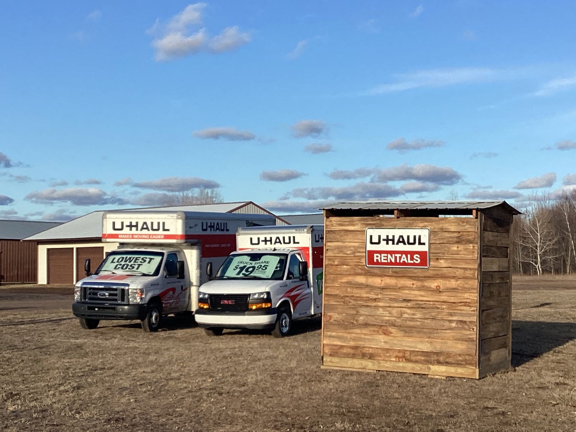 Two u-haul trucks are parked next to a wooden shed in a field.