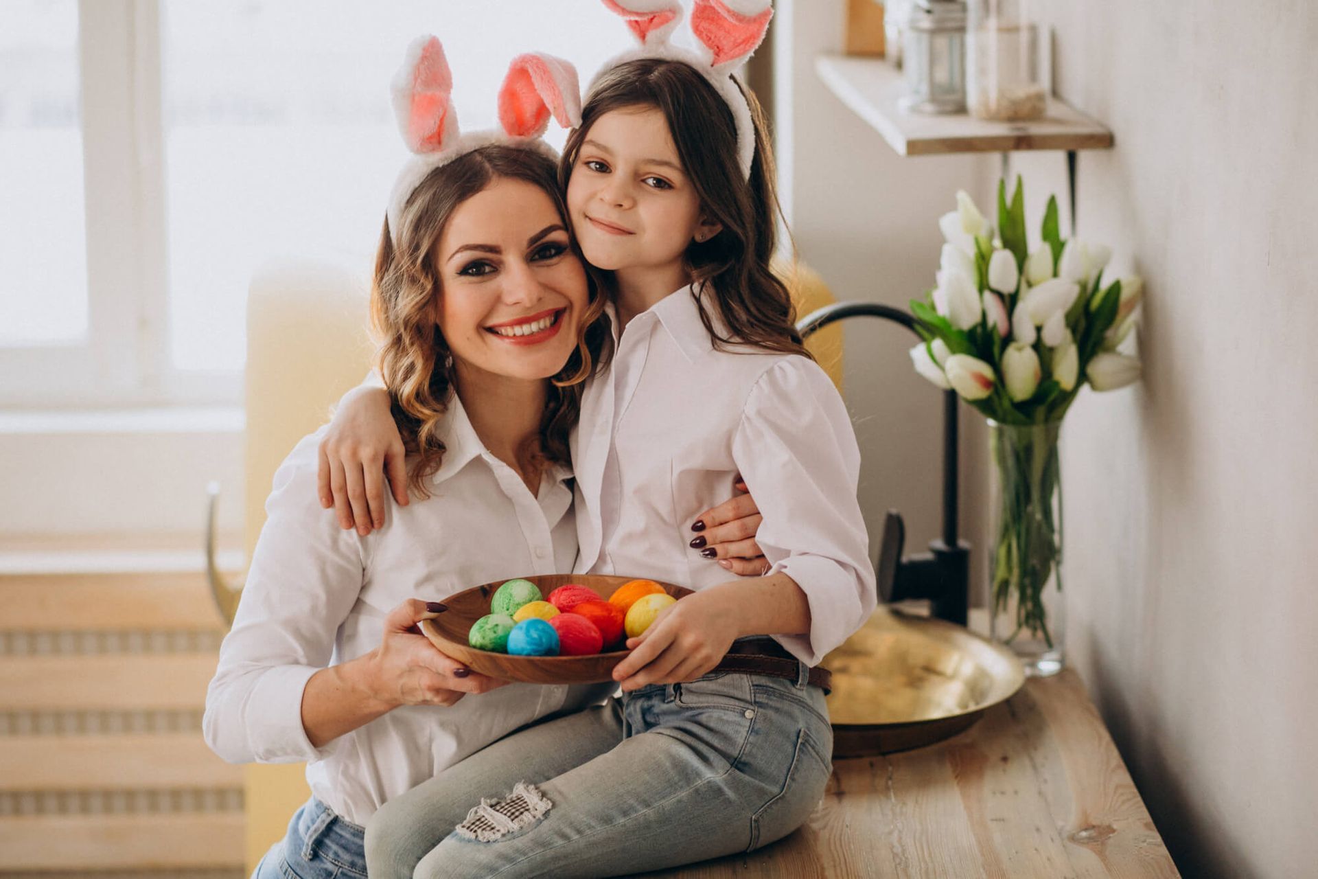 A woman and a little girl are sitting at a table holding a bowl of easter eggs.