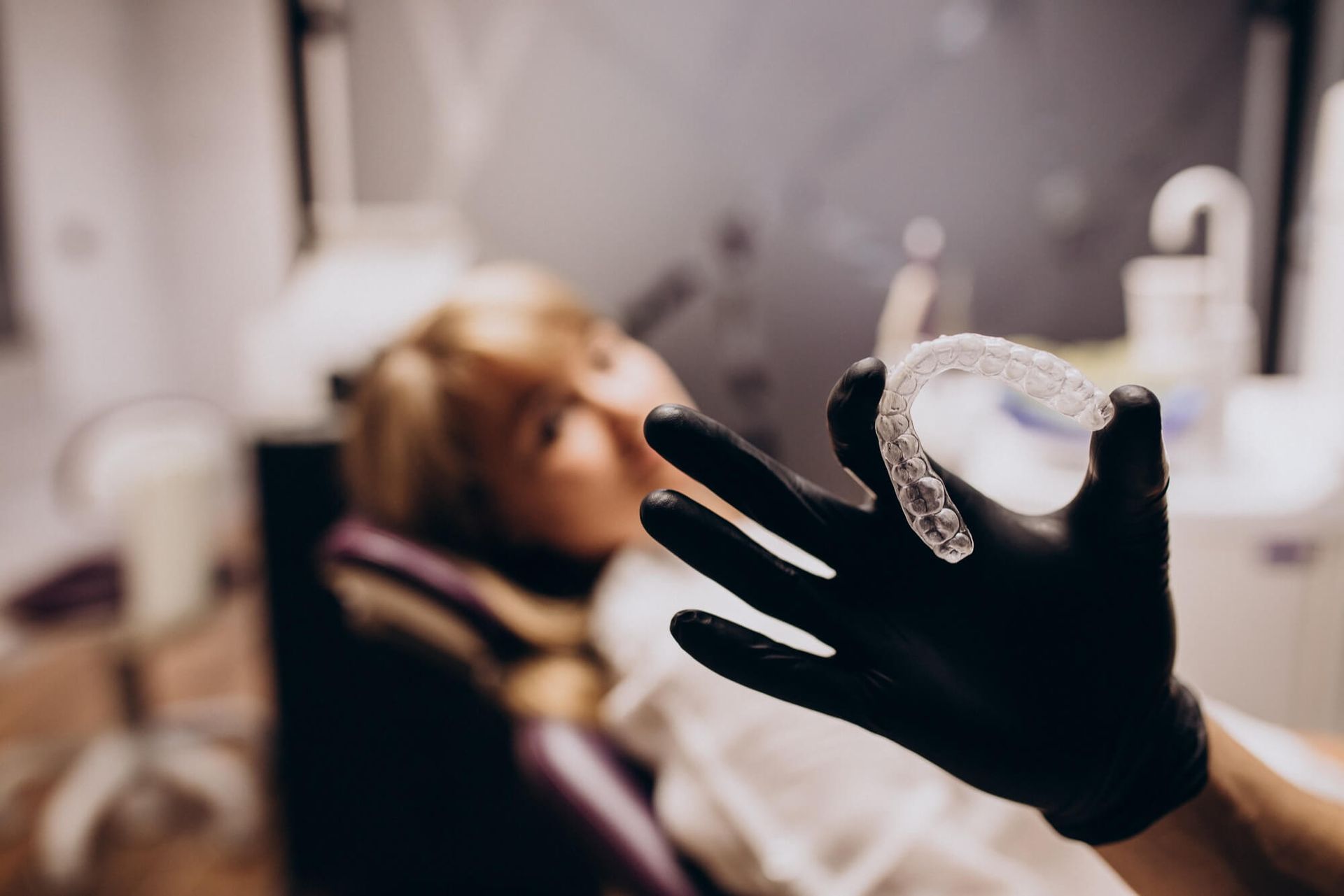 A woman is sitting in a dental chair while a dentist holds a clear brace in his hand.
