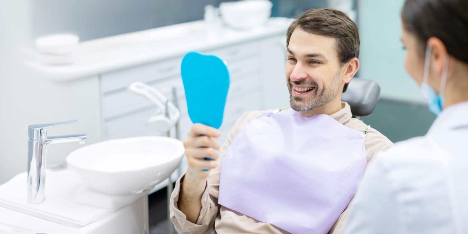A man is sitting in a dental chair looking at his teeth in a mirror.