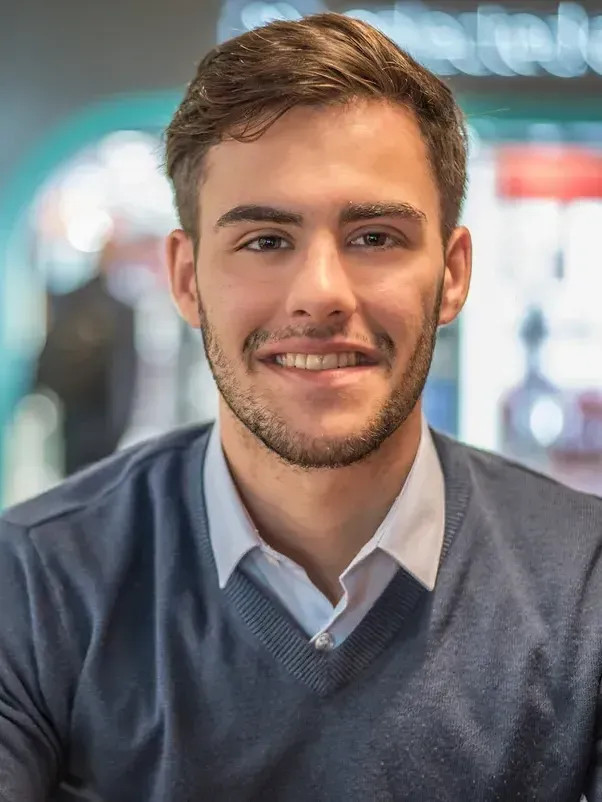 A young man with a beard wearing a blue sweater and white shirt is smiling for the camera.