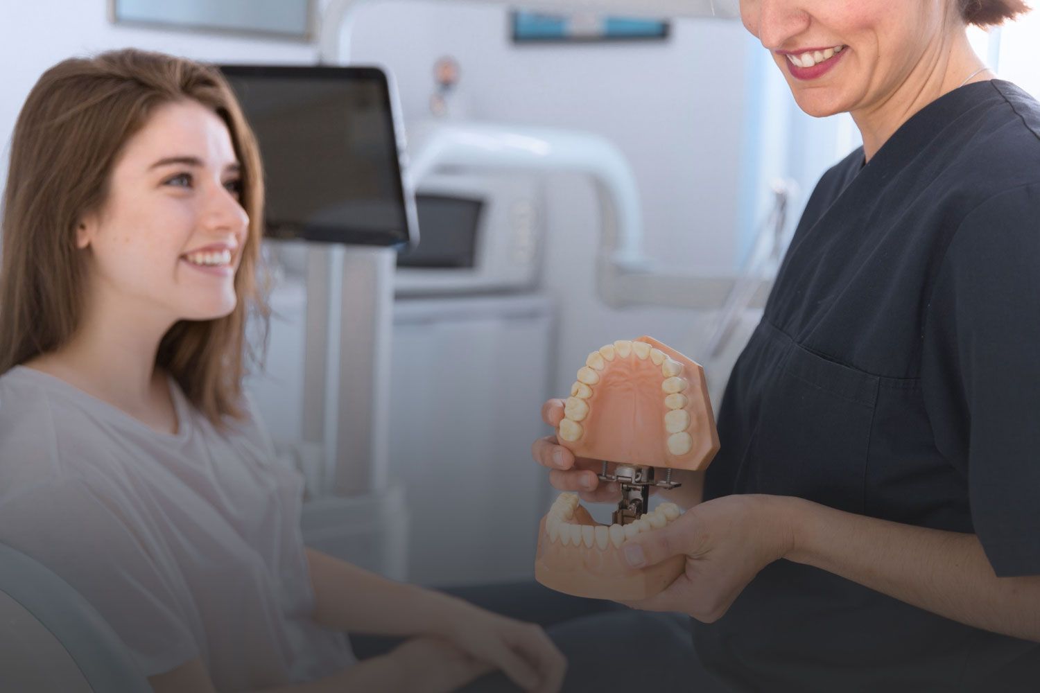 A woman is sitting in a dental chair while a dentist holds a model of her teeth.