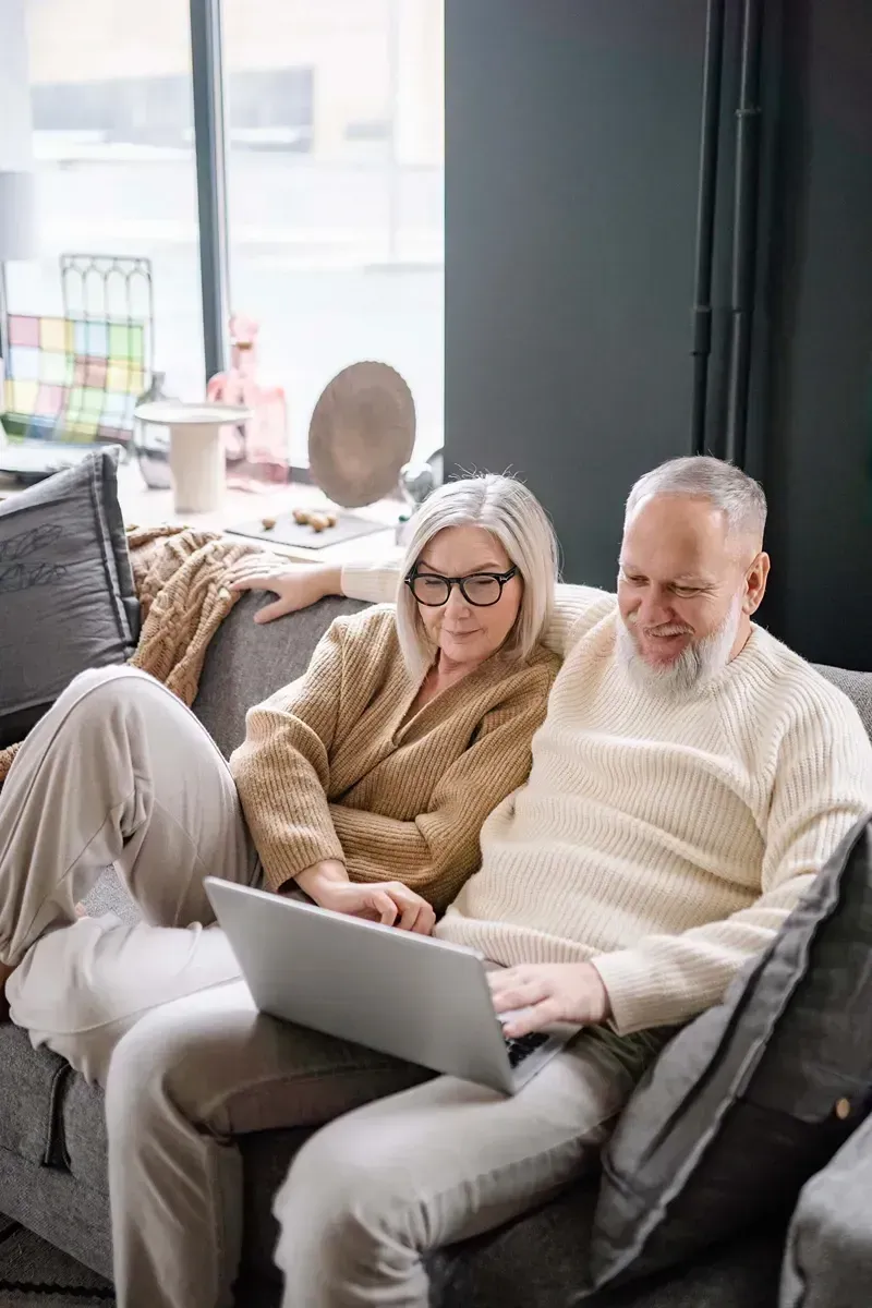 An elderly couple is sitting on a couch using a laptop computer.