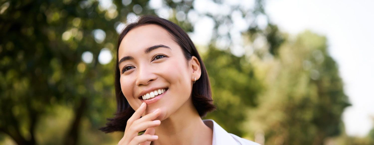 A woman is smiling with her hand on her chin in a park.