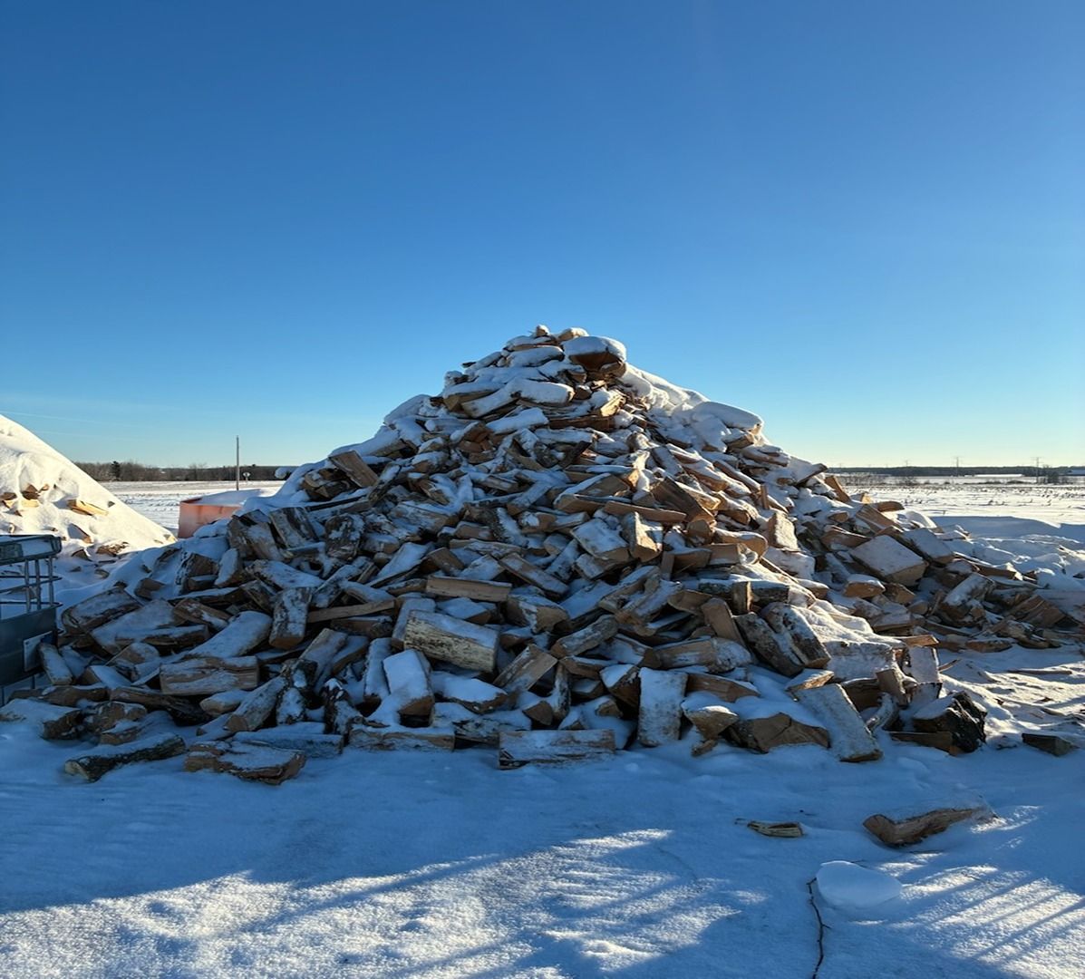 Snow-covered pile of lumber or debris in an open field under a clear blue sky