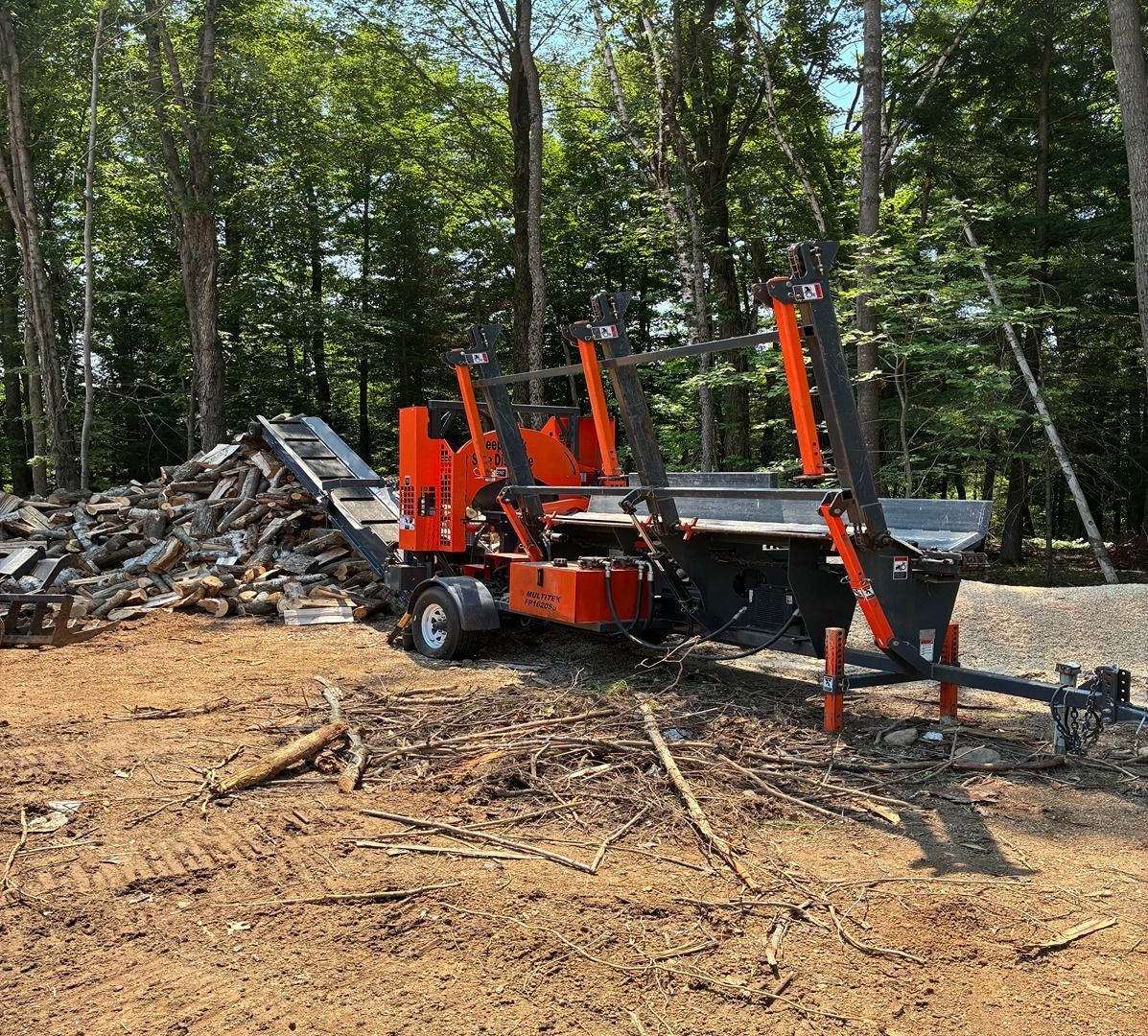 Orange log splitter in a wooded clearing beside a pile of cut logs and a trailer hitch.