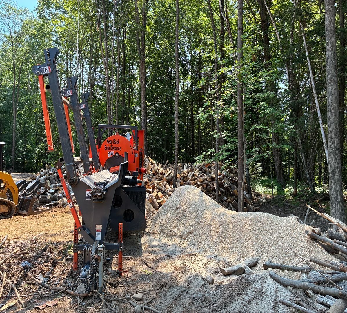 Orange wood chipper in a wooded area beside a large pile of sawdust and cut logs