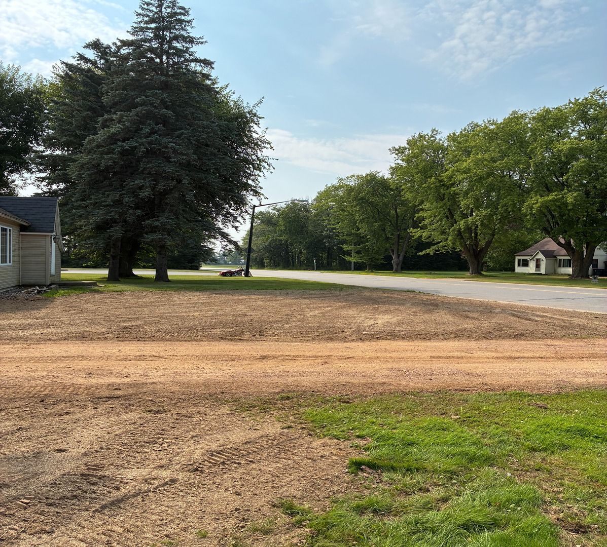 Empty dirt lot beside a road, with green trees and a small house under a clear sky.