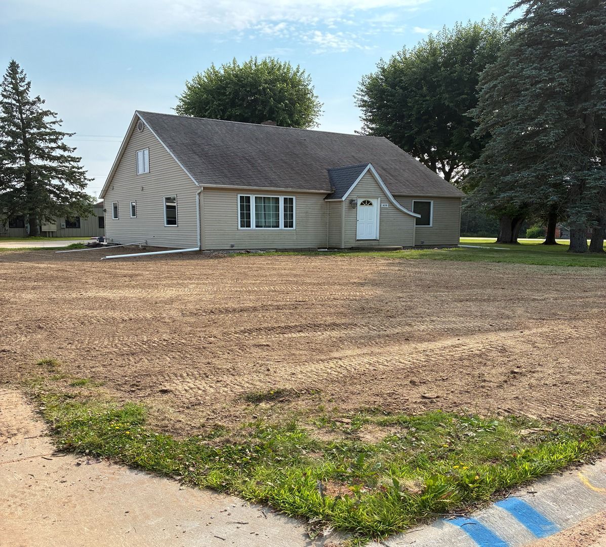Small beige house with a white door, set behind a bare yard with trees and a partly visible sidewalk.