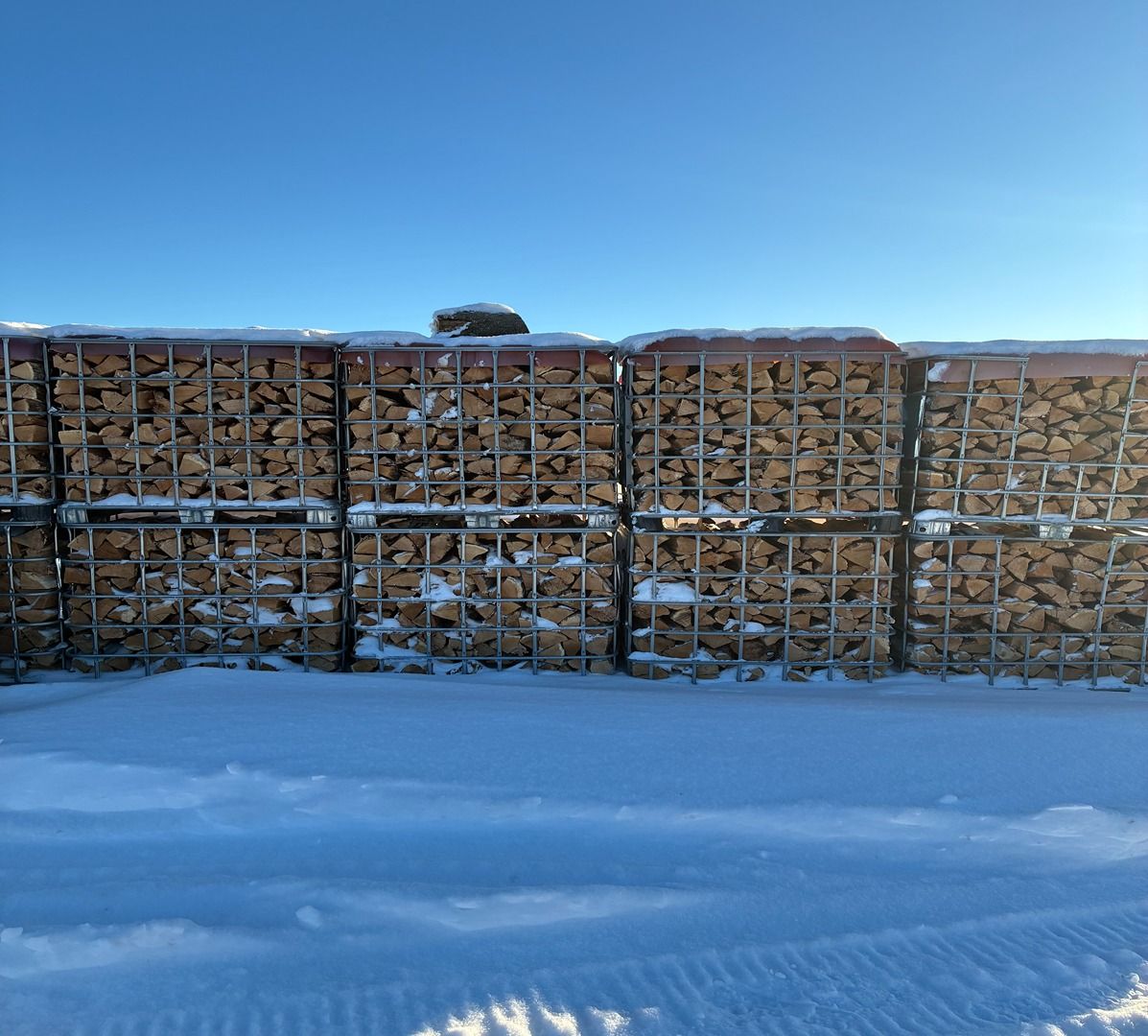 Stacked wood in crates behind a snowy field under a clear blue sky