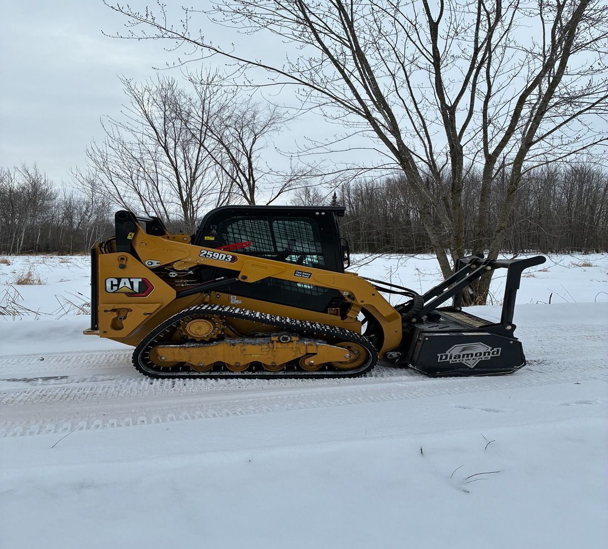 Yellow CAT snow loader with plow attachment clearing snow on a road near bare trees.