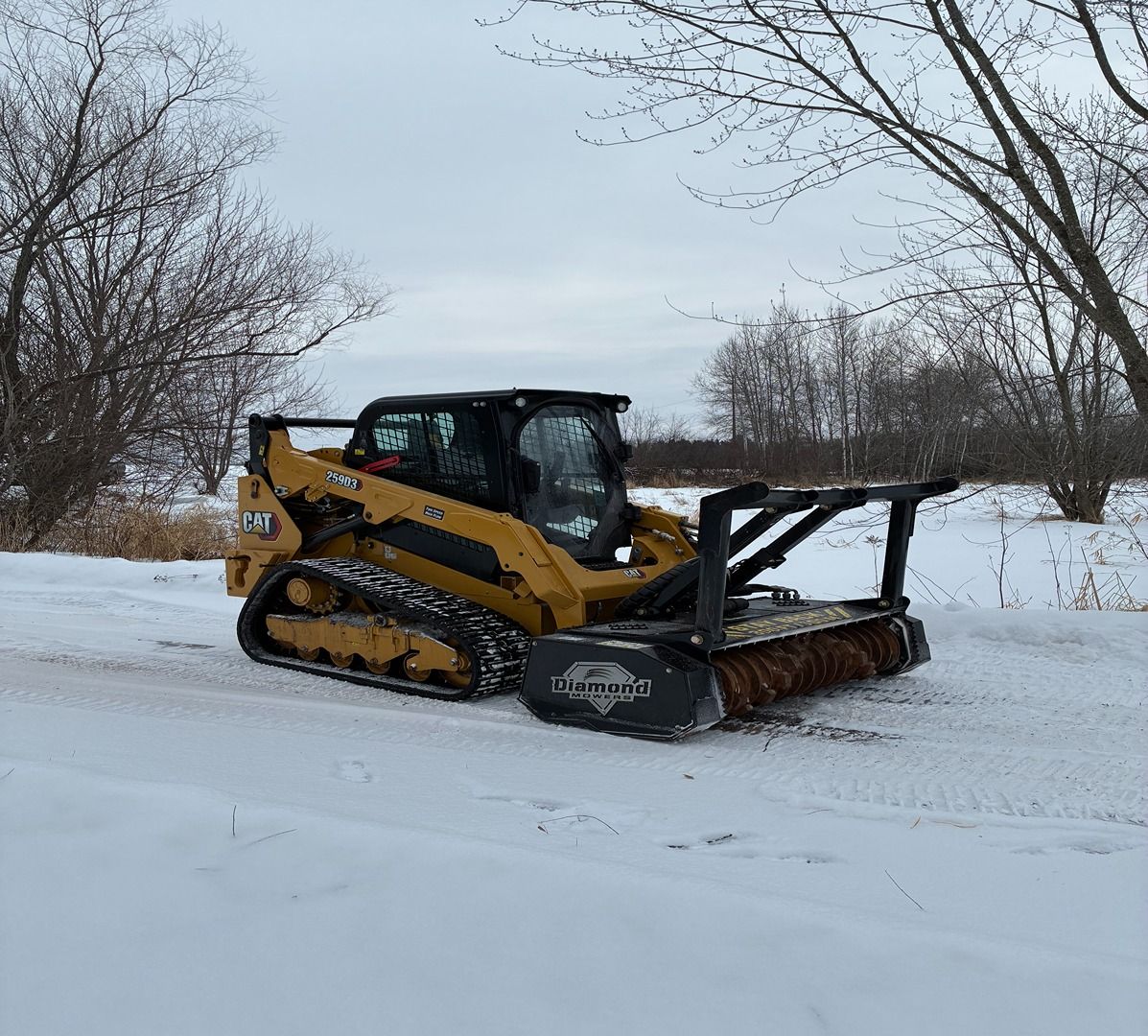 Yellow compact track loader pushing snow on a snowy road with bare trees in the background