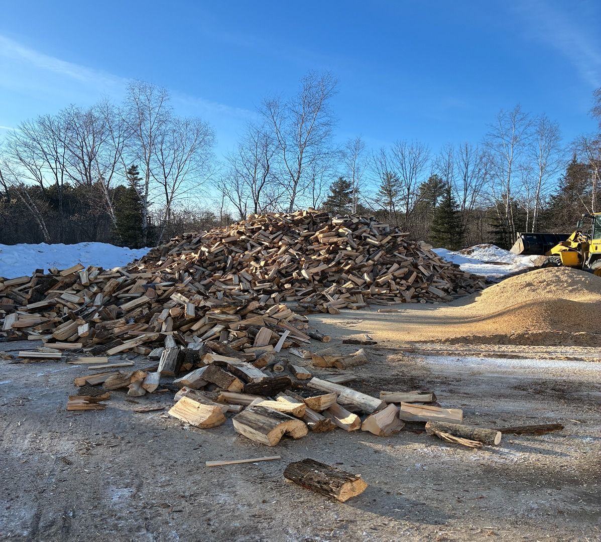 Large pile of wood chips and logs in a yard under a blue sky, with a yellow loader on the right.