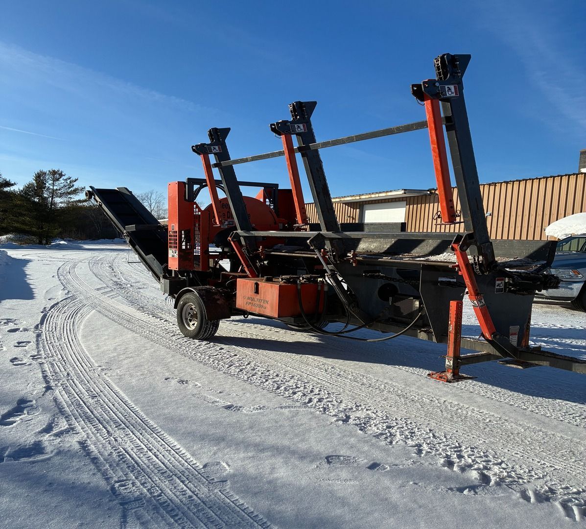 Red snow-removal machine with black arms on a snowy road under a clear blue sky.
