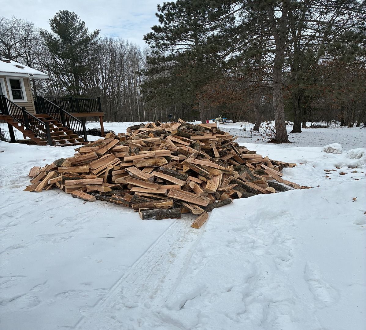 Large pile of split firewood on snowy ground beside a house and trees