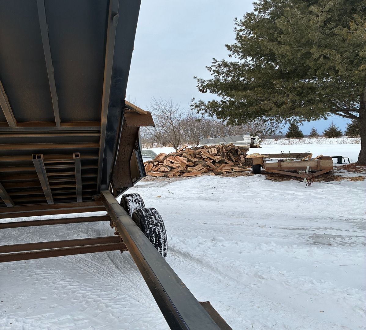Snowy yard with a woodpile, picnic table, and large tree beside a house deck.