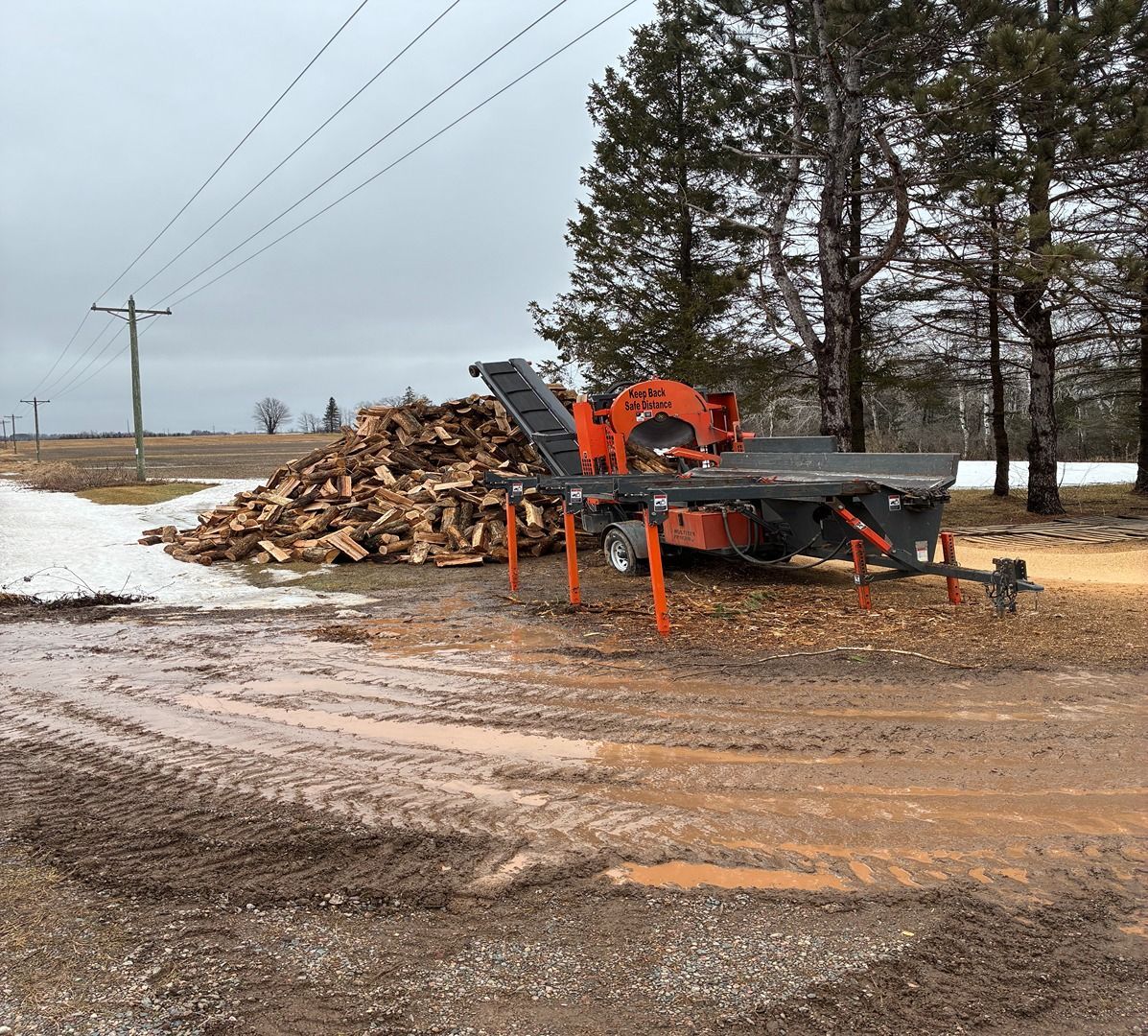 Orange wood splitter beside a large log pile on a muddy roadside in winter