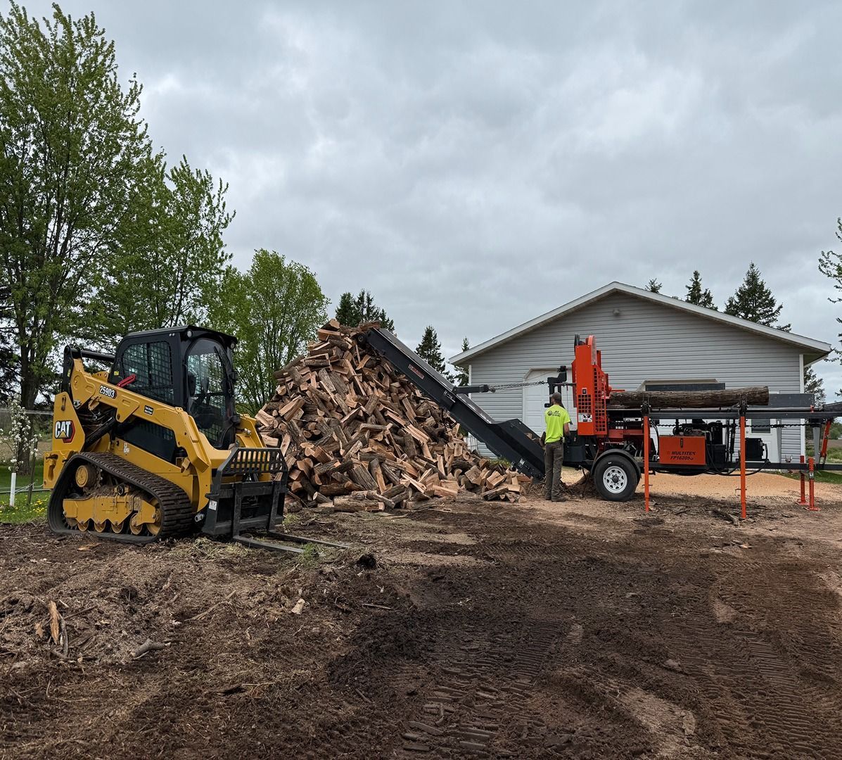 Yellow skid steer and orange excavator moving dirt beside a house under cloudy skies