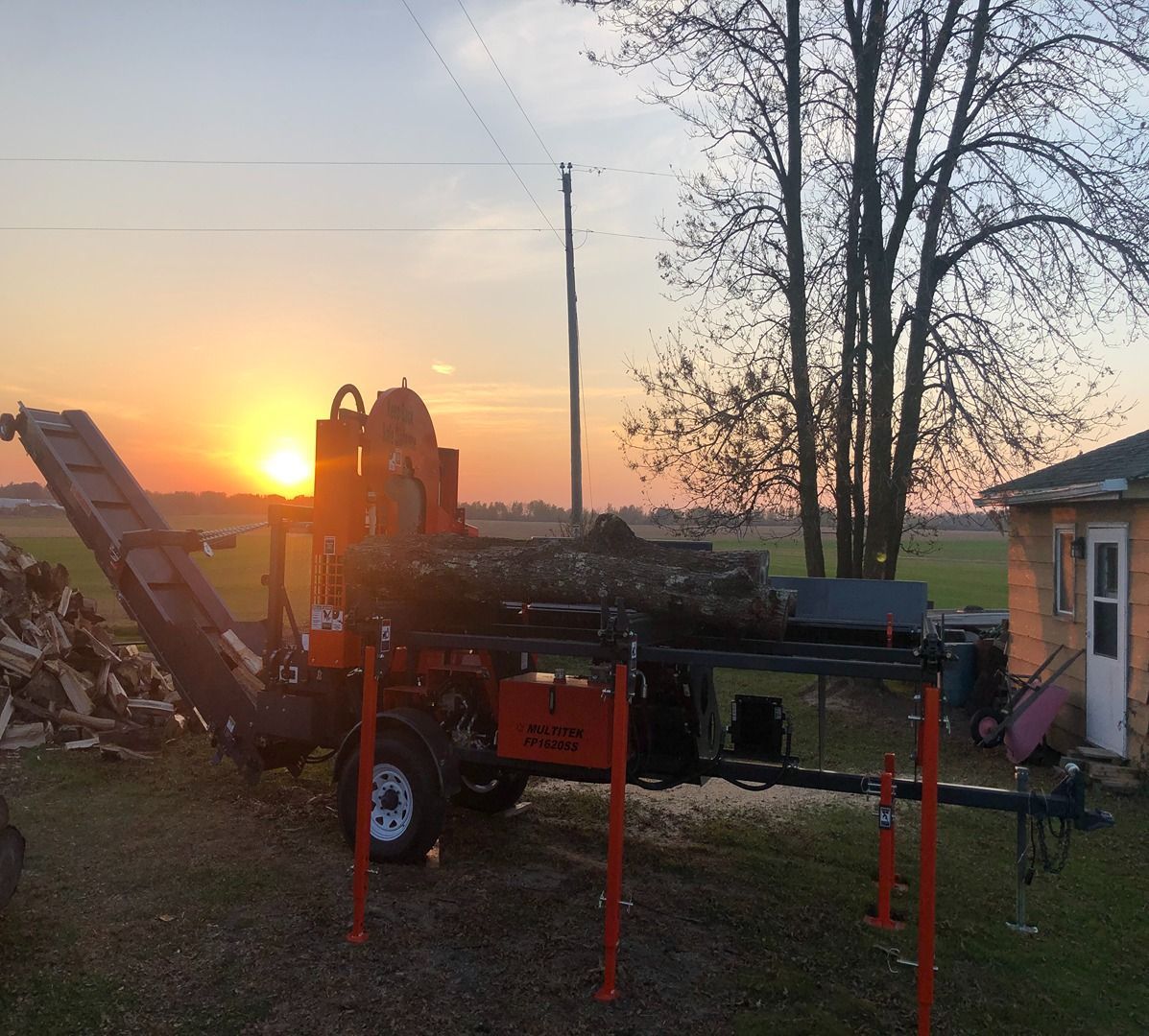 Sunset over a yard with farm equipment, red machinery, and a small house beside bare trees