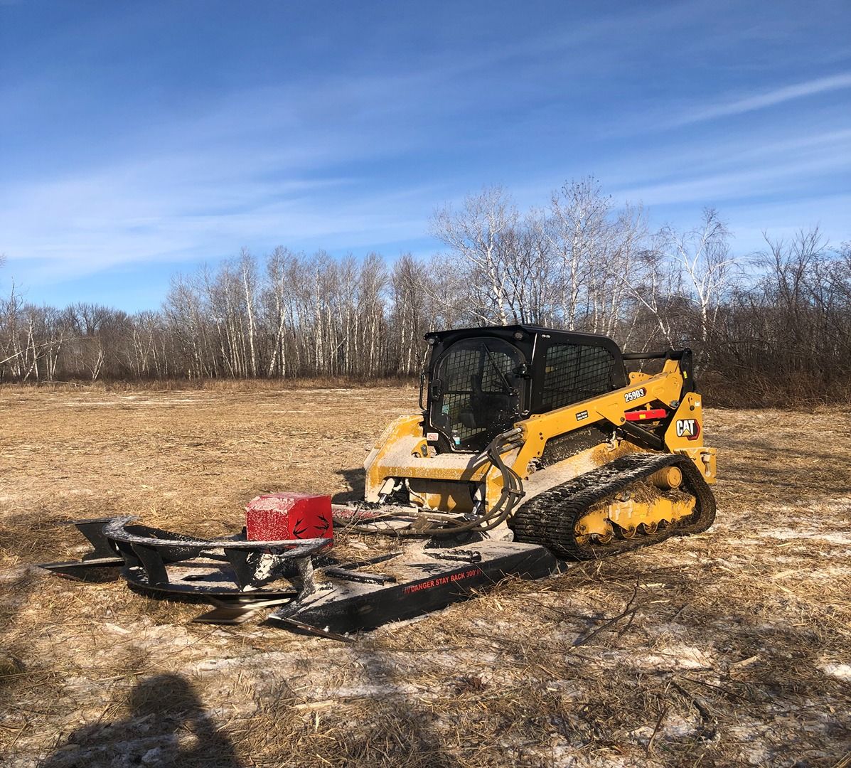 Yellow track loader in a field, pushing a red attachment under a blue sky