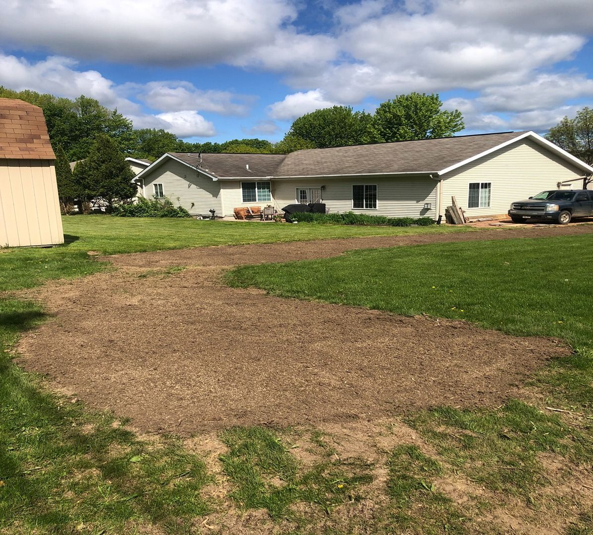 Backyard with a large dirt patch, green grass, and a white house under a partly cloudy sky