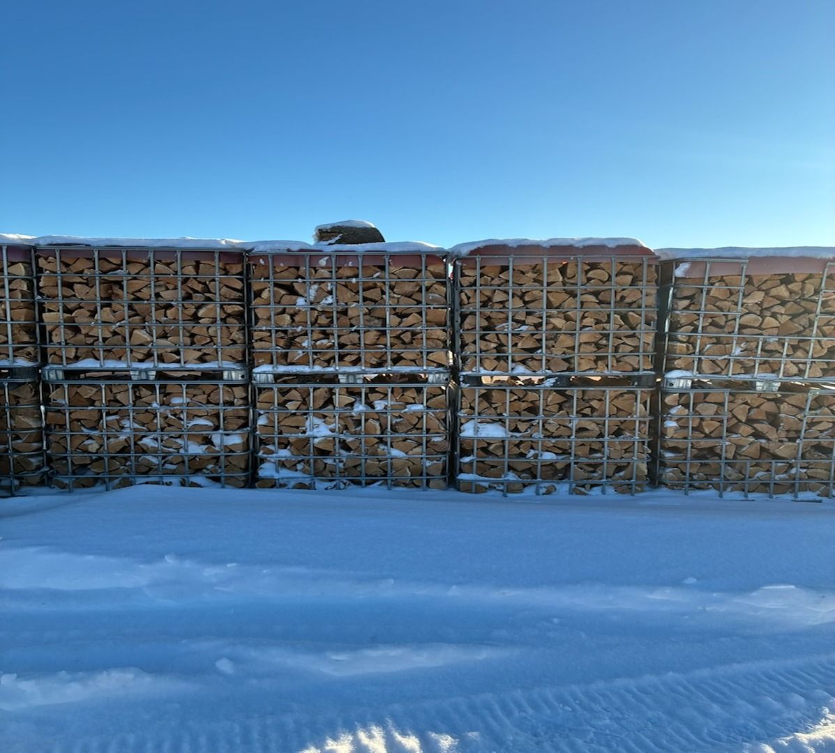 Snow-covered woodpile stacked behind wire fencing under a clear blue sky.