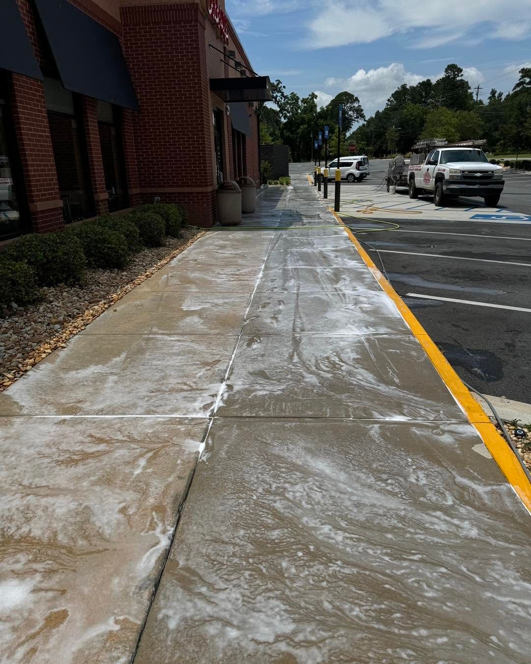 Sidewalk with white residue beside a brick building, next to a parking lot with cars under a blue sky.