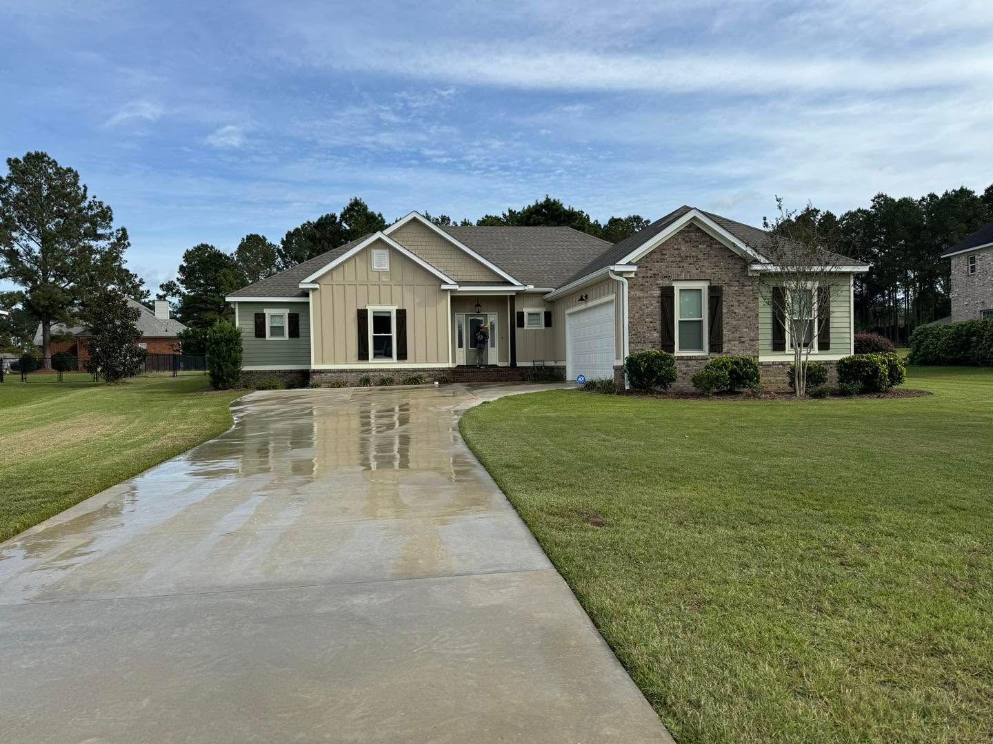 Single-story beige house with stone accents, wet driveway, and green lawn under a cloudy sky