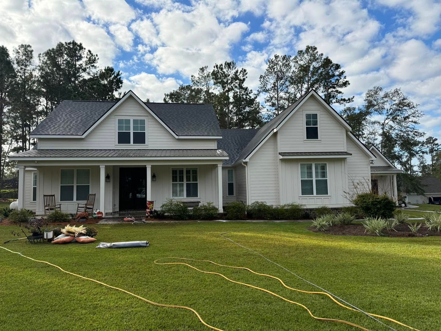 Single-story white house with gray roof and front porch, lawn sprinkler hoses across the yard, trees behind