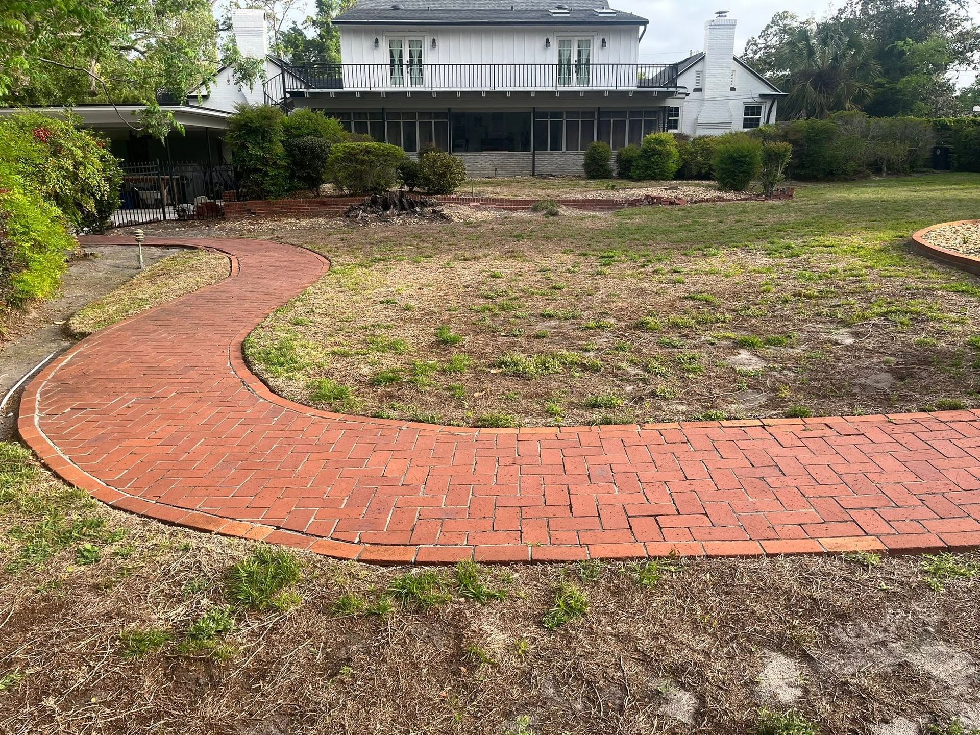 Curved red brick path through a dry yard leading to a white house with a porch and trees in front