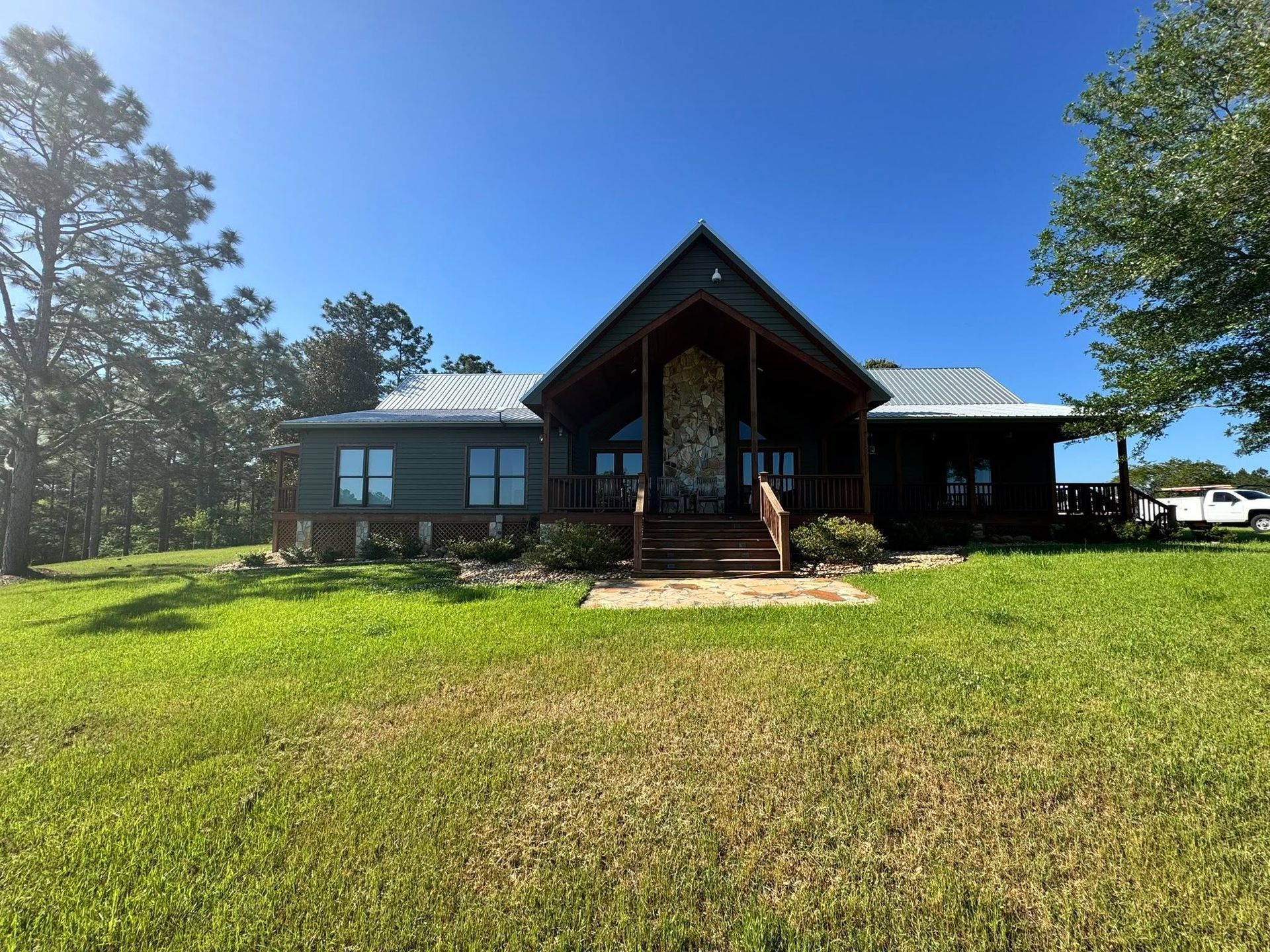 Single-story dark house with a front porch, centered on a sunny green lawn under a blue sky