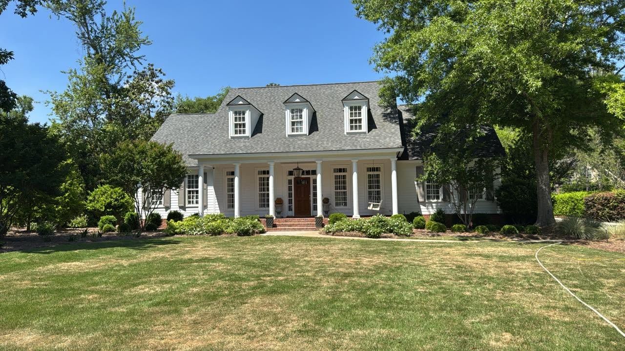 White two-story house with dormer windows, front porch, and a large grassy lawn under sunny skies
