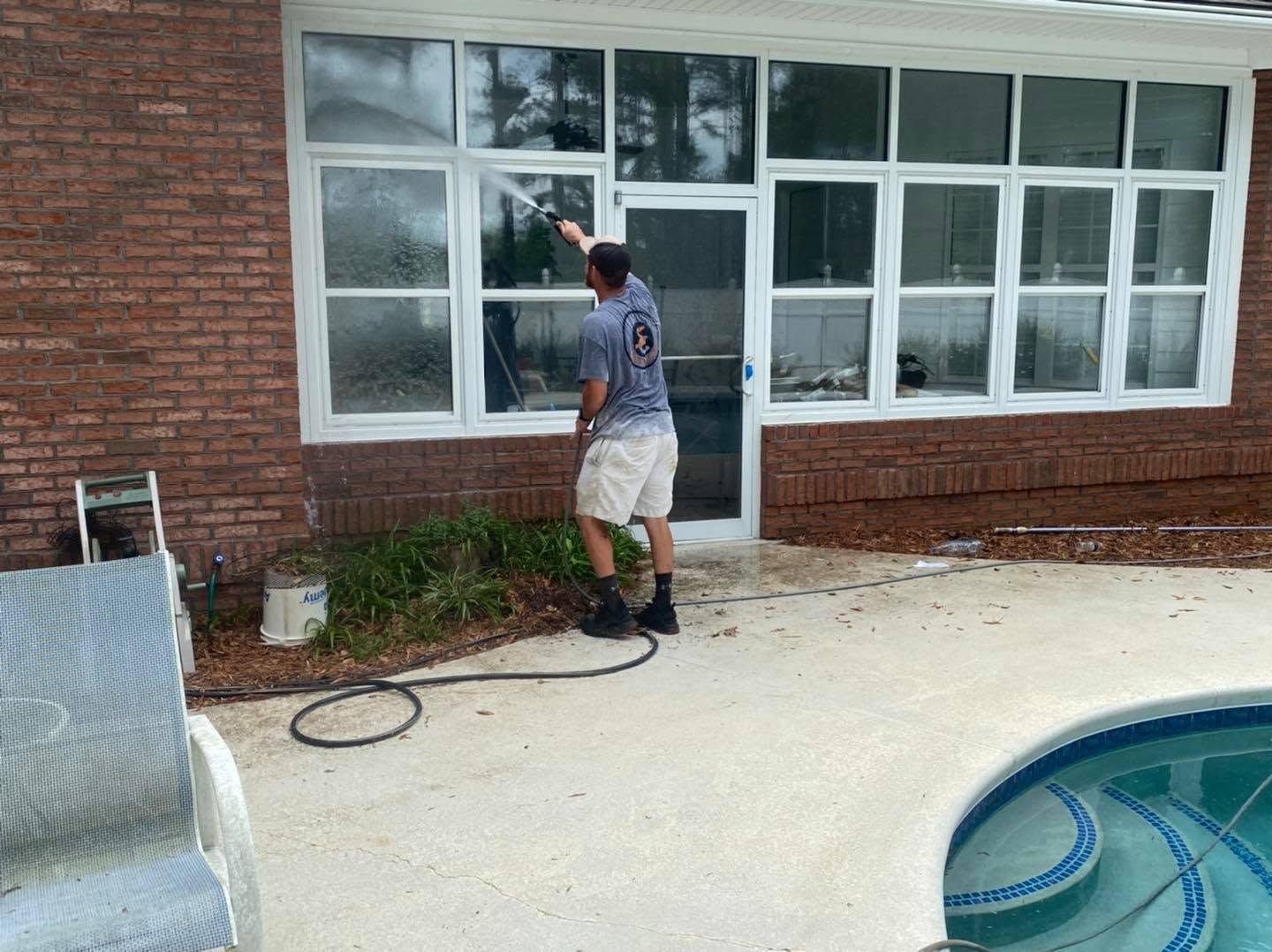 Person pressure washes the exterior wall of a brick house beside a swimming pool.