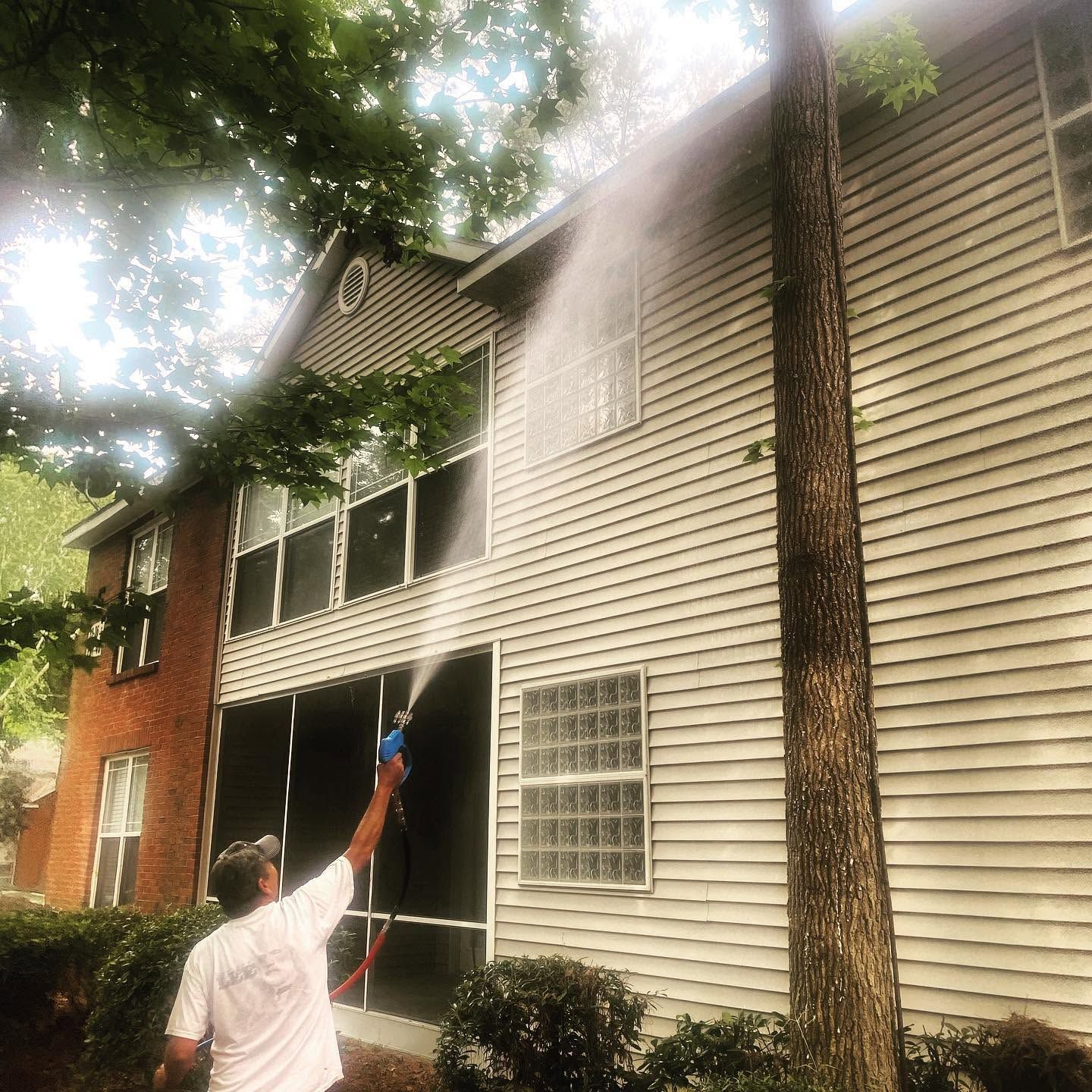 Worker sprays the exterior of a two-story house with a pressure washer under trees.