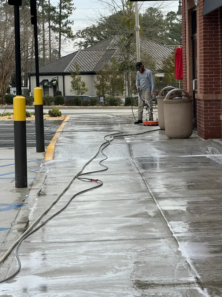 Worker pressure-washing a wet concrete walkway beside a brick building on a rainy day
