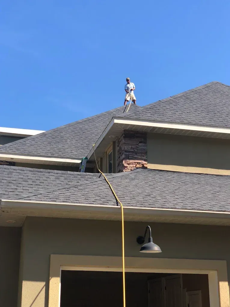 Worker on a roof, attached to a yellow rope, with a ladder below and blue sky overhead