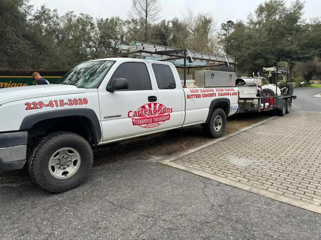 White work truck towing a trailer with a red logo, parked on a gravel road near trees.