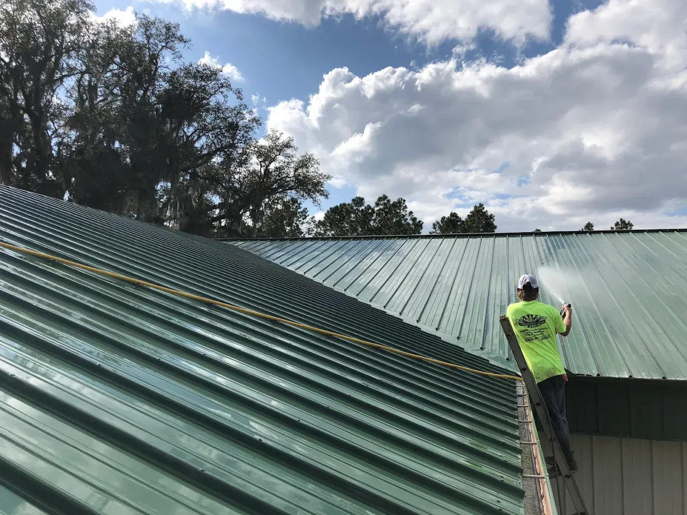 Worker on green metal roof spraying water under cloudy sky