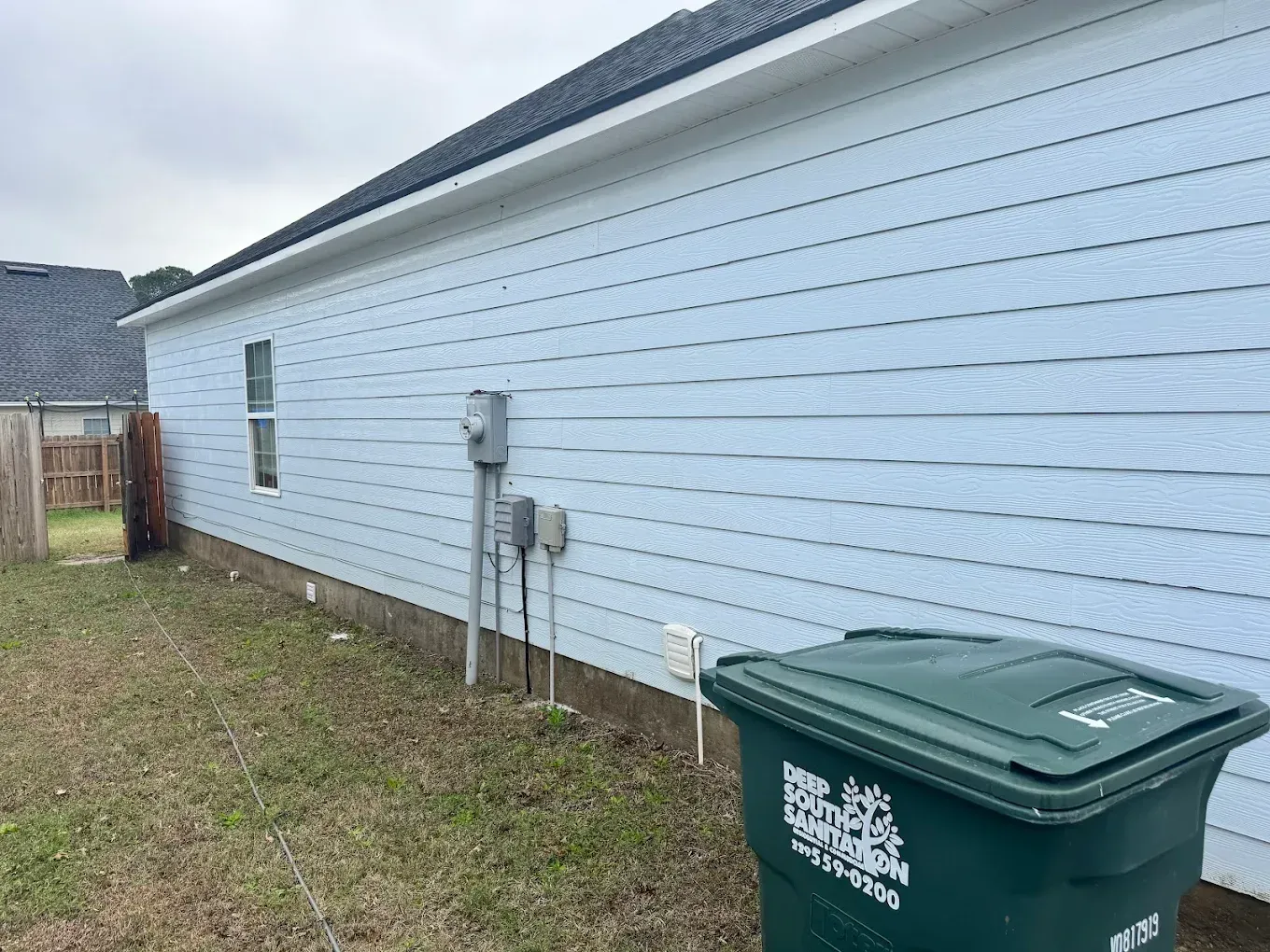 White house side wall with utility meters and a green trash bin beside a grassy yard.
