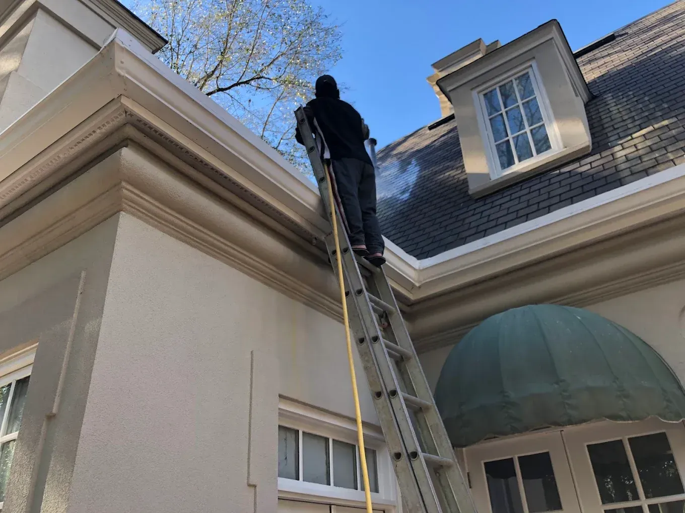 Person on a ladder cleaning a house gutter beside a dormer window and green awning