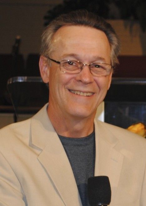 A man wearing glasses and a jacket is smiling and holding a microphone