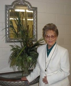 An elderly woman in a white suit is standing next to a vase of flowers.