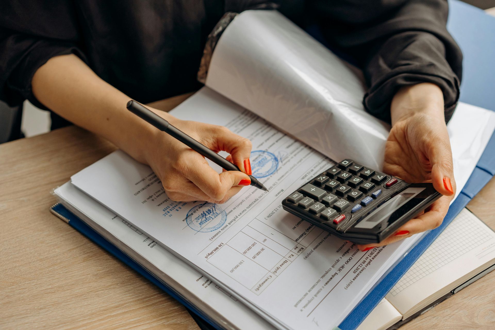 Person with red nails writing on a document with a calculator on the side. Papers and a notepad are on a desk.