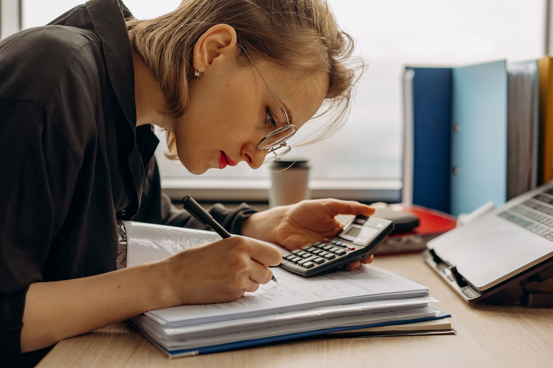 Person writing on a notepad while using a calculator at a desk with books and a laptop