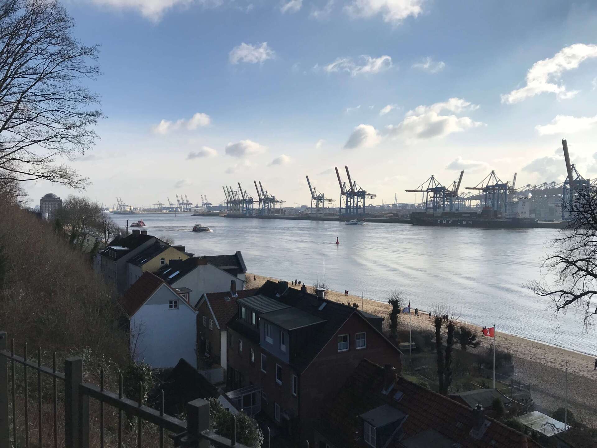 Blick auf den Hamburger Hafen, Fluss mit Schiffskränen und Gebäuden unter bewölktem Himmel.