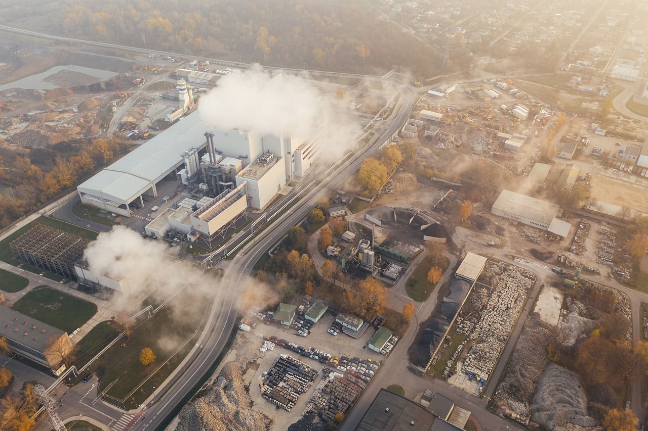 Eine Fabrik stößt weißen Rauch in einen dunstigen Himmel aus, umgeben von Industriegebäuden und Trümmern.