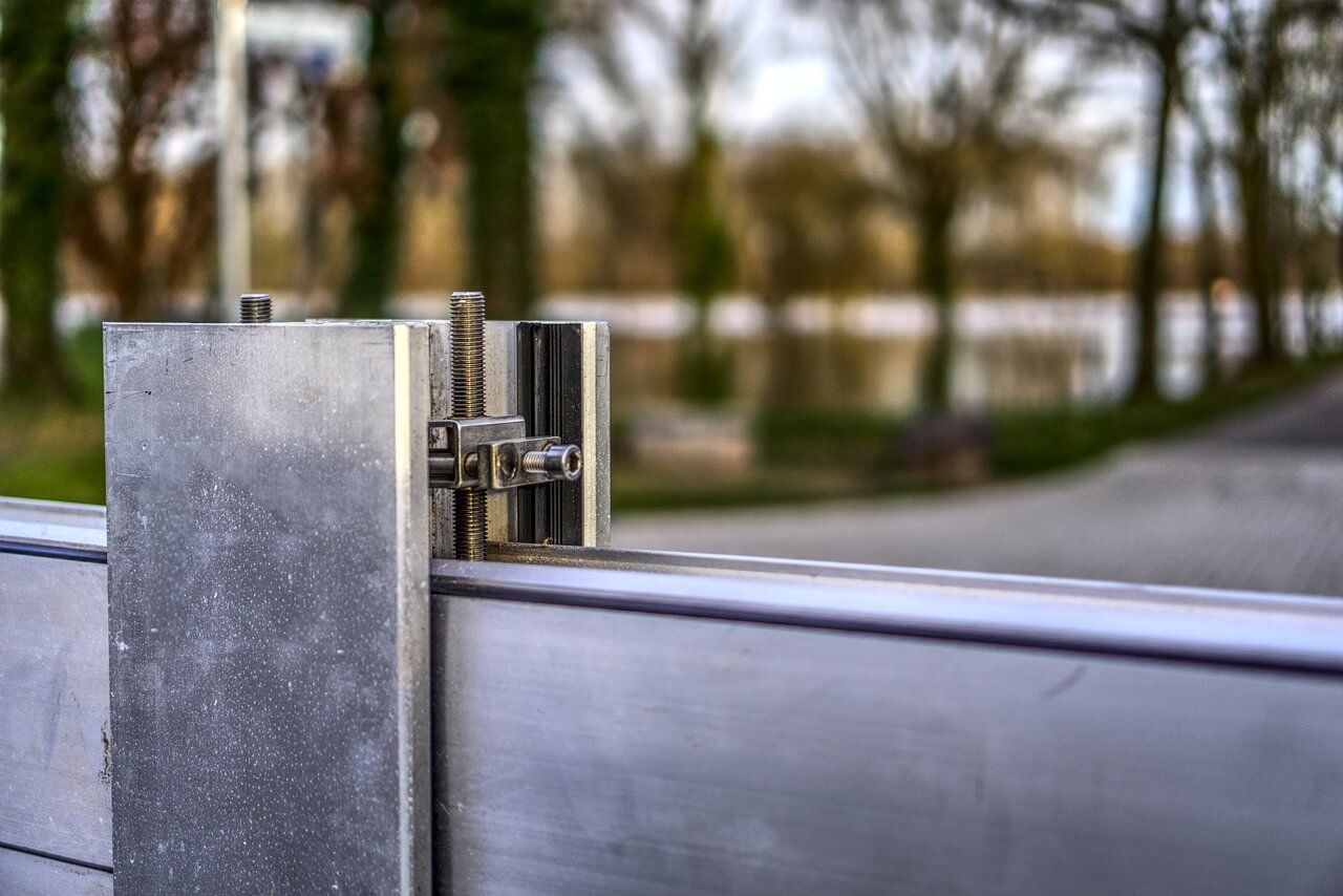 Metal flood barrier detail, gray and silver, with trees and road in the blurred background.