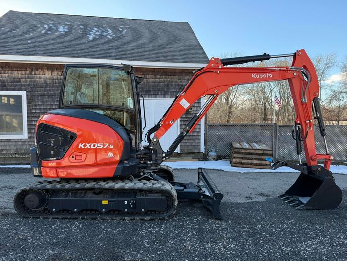 Red compact excavator with bucket parked on gravel beside a weathered wooden building.